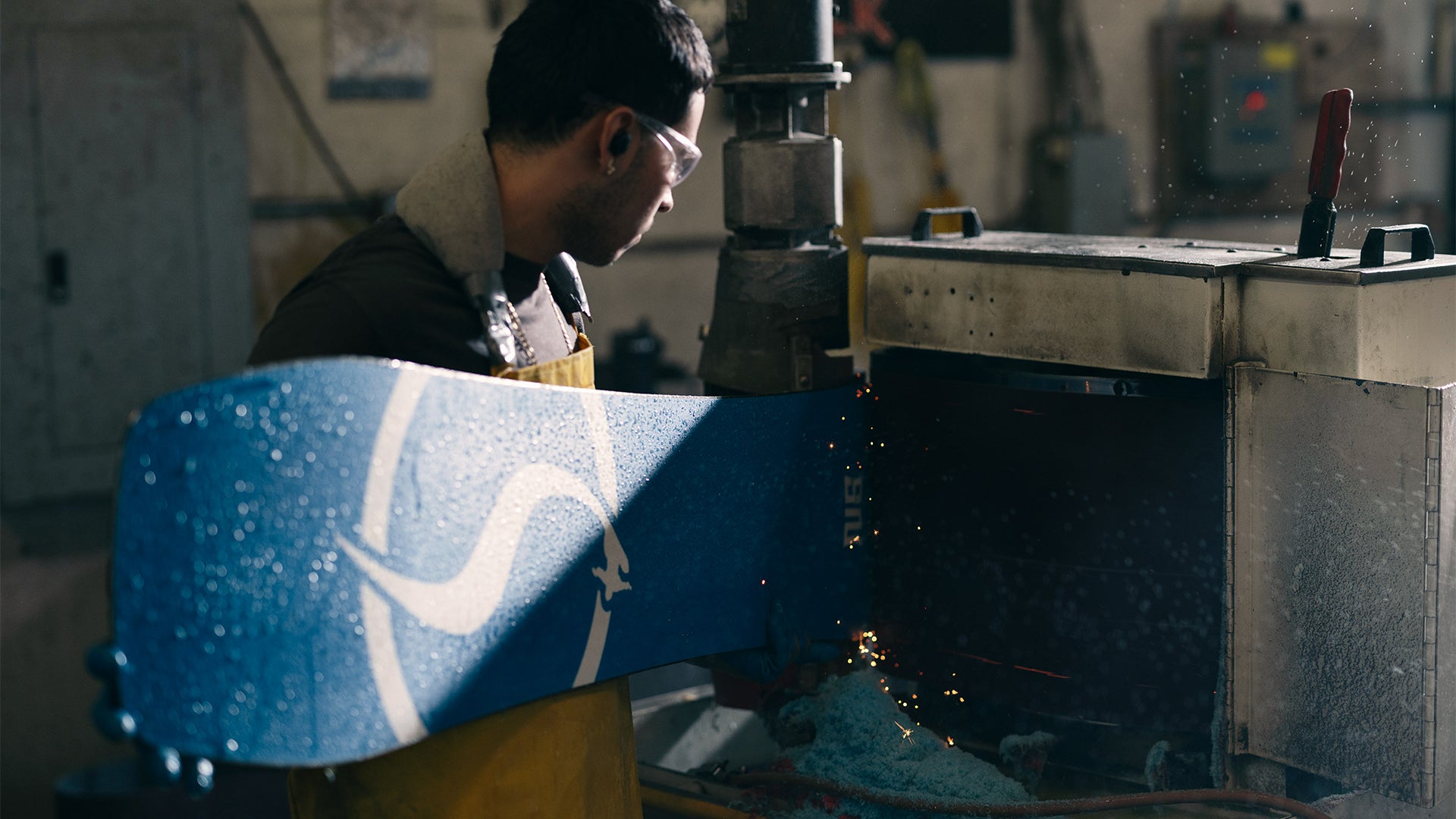 A snowboarder in a workshop setting, holding a blue snowboard with a white graphic design while a machine cuts it, with sparks flying.